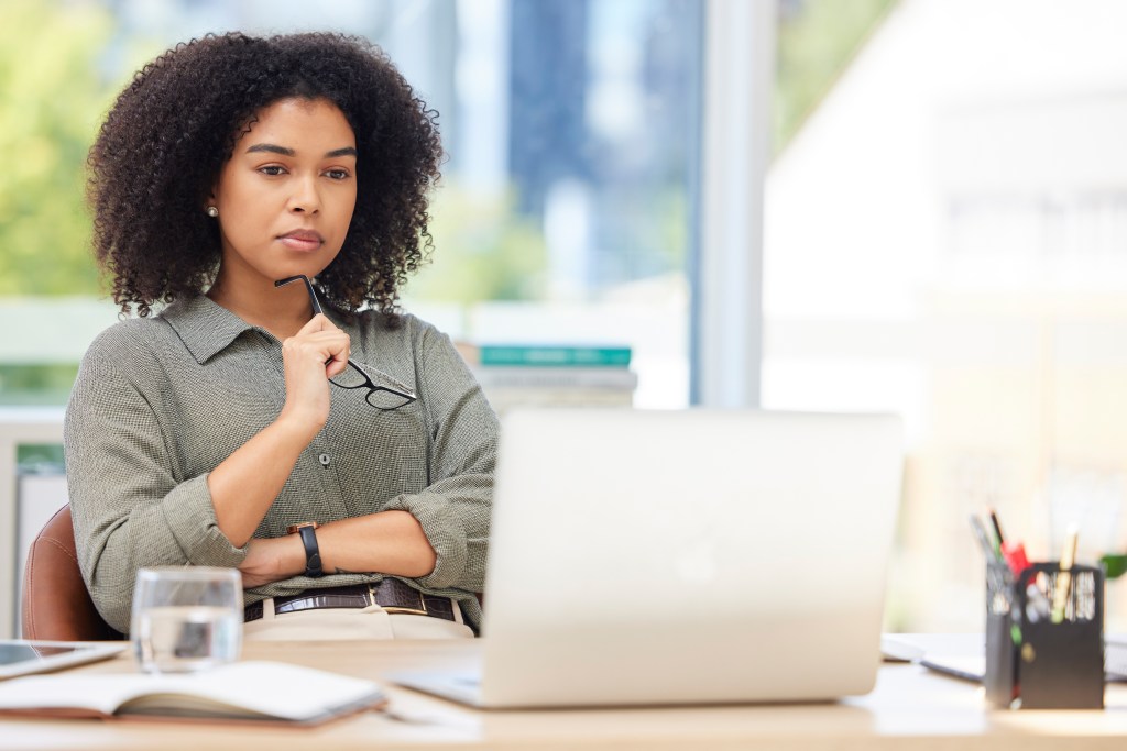 A woman with curly hair sitting at a desk, thoughtfully holding glasses, looking at a laptop computer in a well-lit office environment.