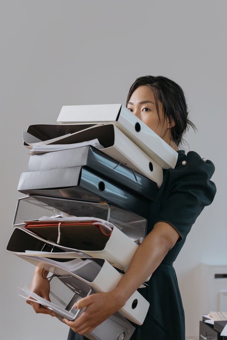 A woman carrying a large stack of assorted organizational binders and files, partially obscured by the binders, in a modern office setting.