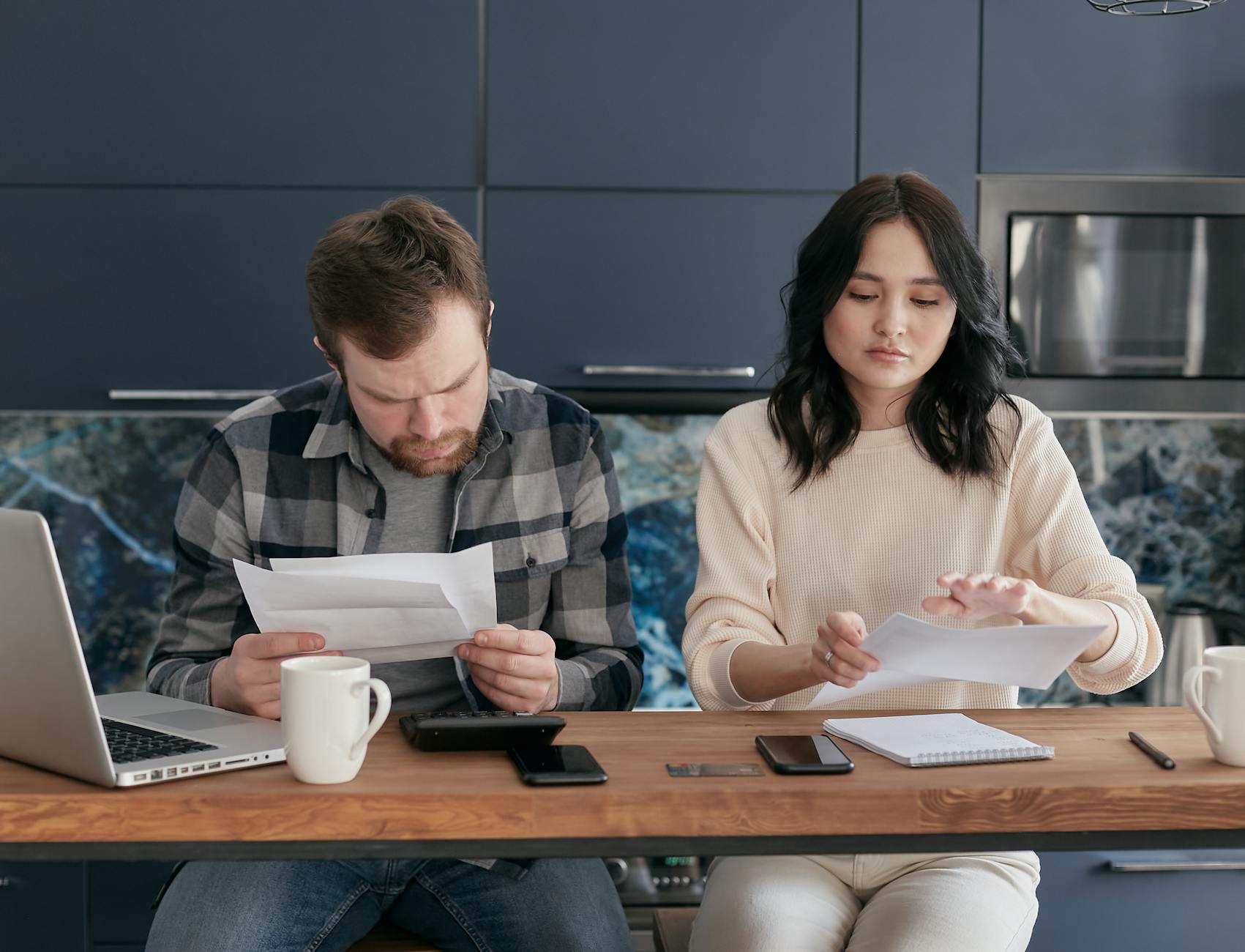 A man and a woman sit at a kitchen table, reviewing documents and notes. The man appears focused on a piece of paper, while the woman is in the process of organizing her papers. A laptop and a coffee mug are on the table.