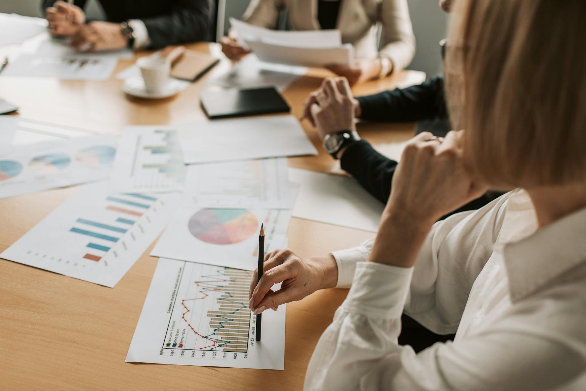 A group of business professionals engaged in a meeting, analyzing various charts and graphs displayed on the table, with one person pointing at a data chart using a pencil.