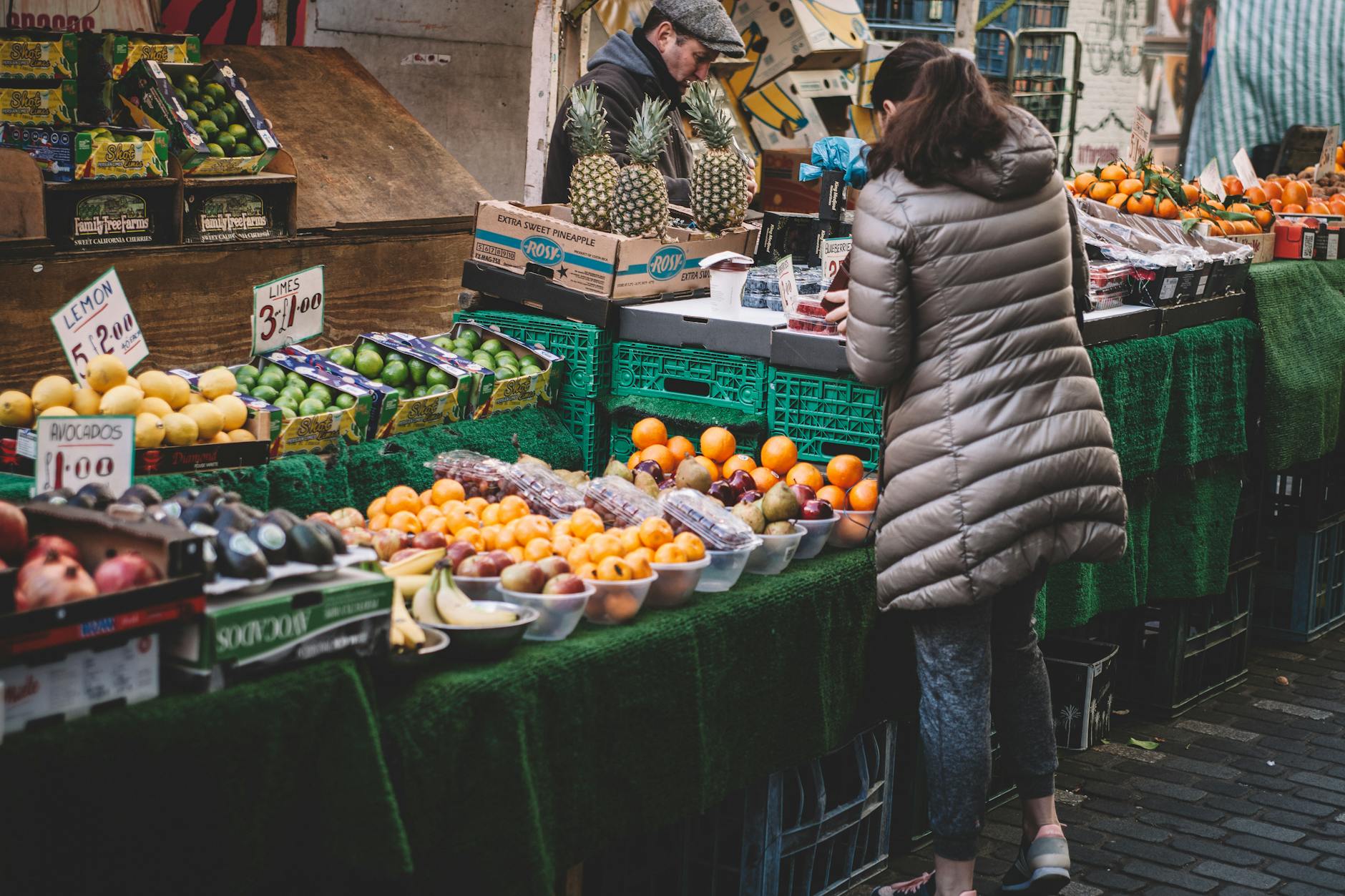 A woman in a puffy gray coat examines fresh produce at an outdoor market stall filled with various fruits, including oranges, avocados, and pineapples, with price tags visible.