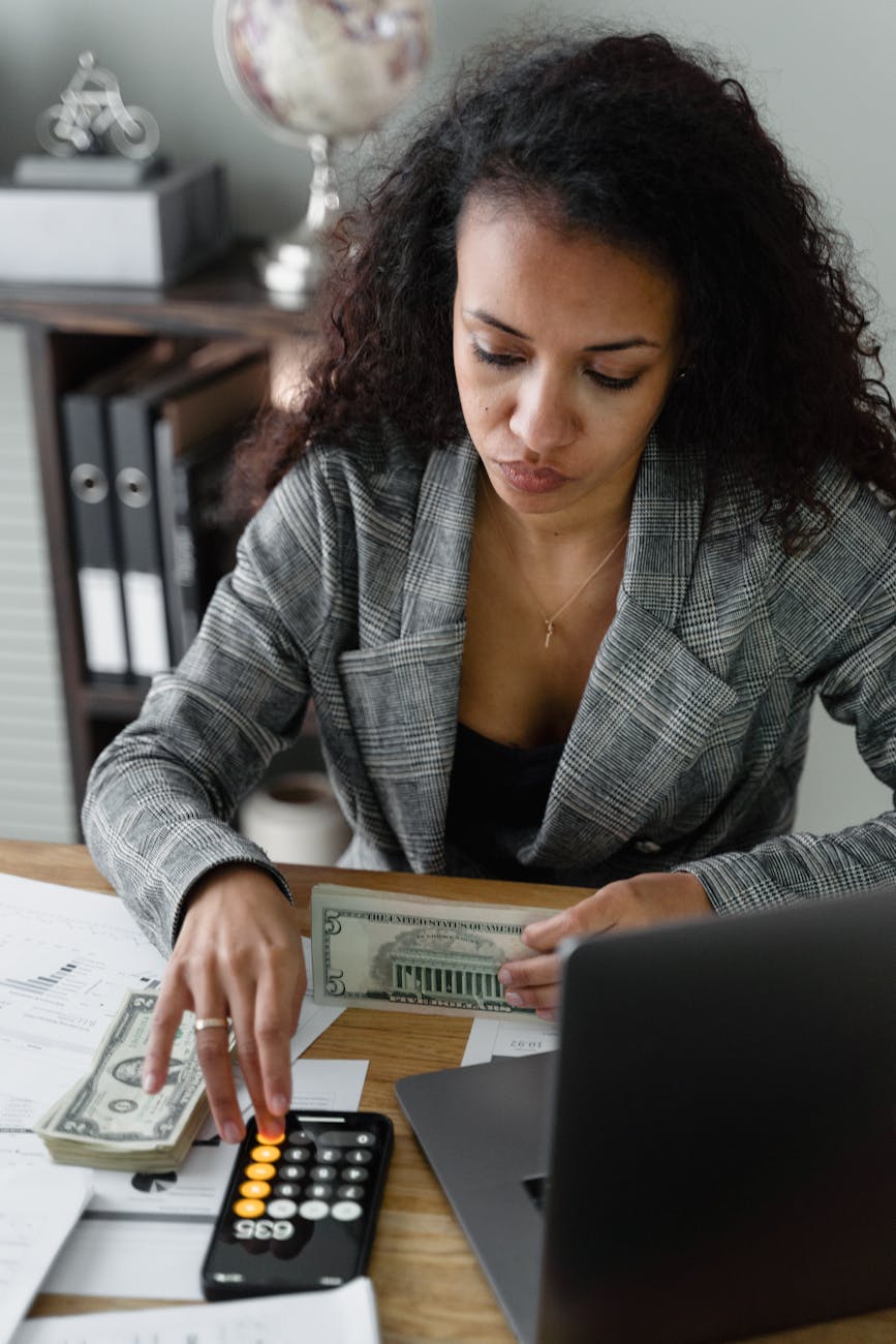 A woman with curly hair is sitting at a desk, reviewing financial documents and holding a five-dollar bill while using a calculator. A laptop and stacks of cash are also on the desk.