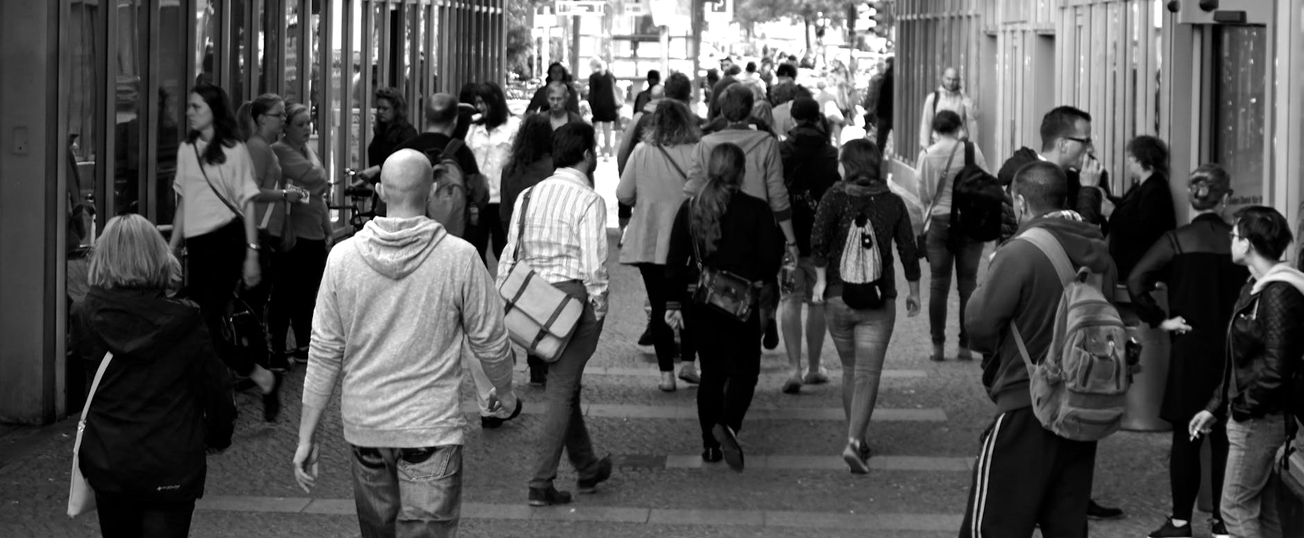 A busy urban street scene in black and white, featuring a diverse group of people walking and interacting. Some individuals are seen walking towards the camera, while others are standing in conversation or observing their surroundings.
