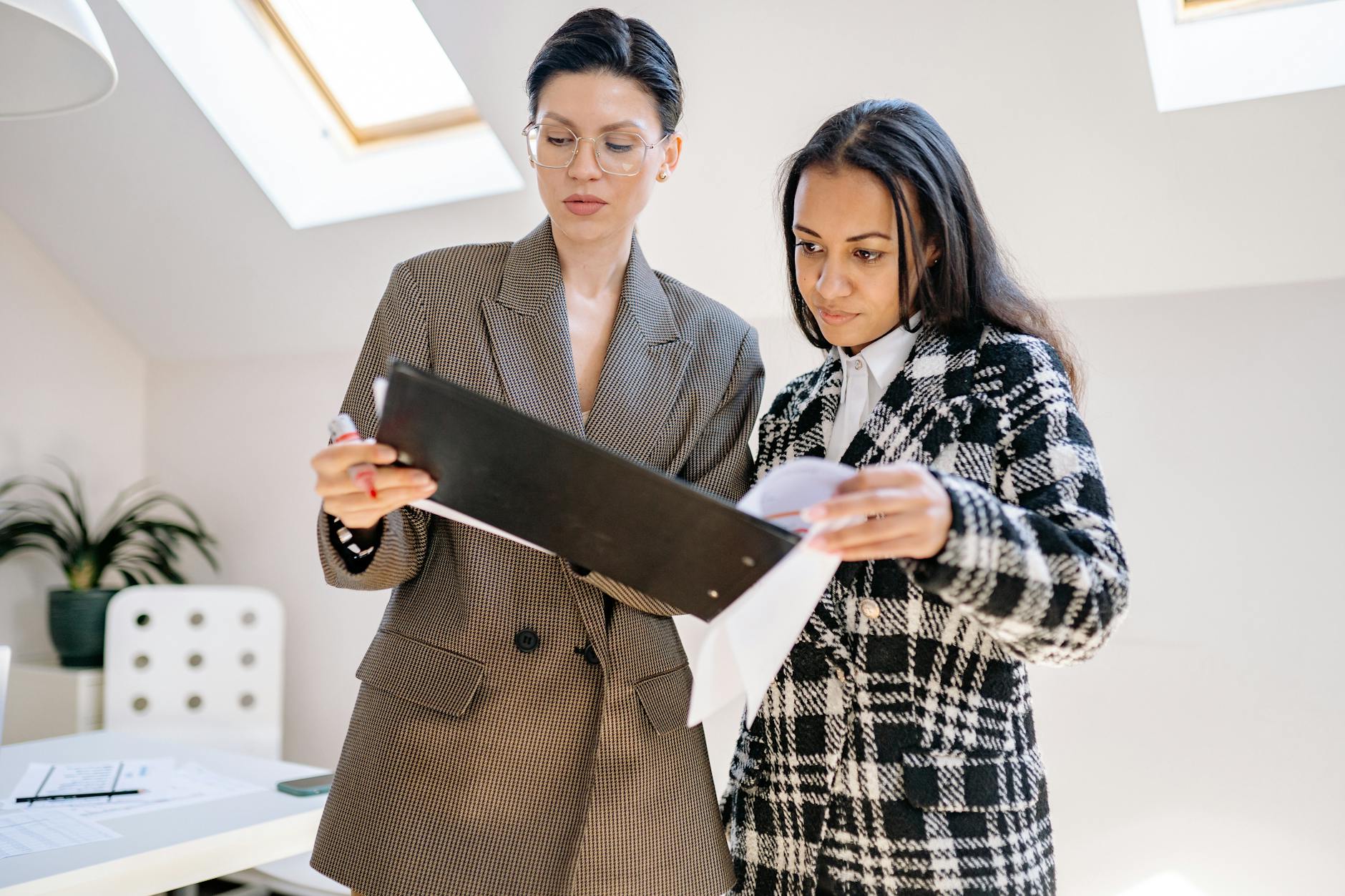 Two professional women discussing documents in an office setting, one holding a clipboard and the other examining papers.